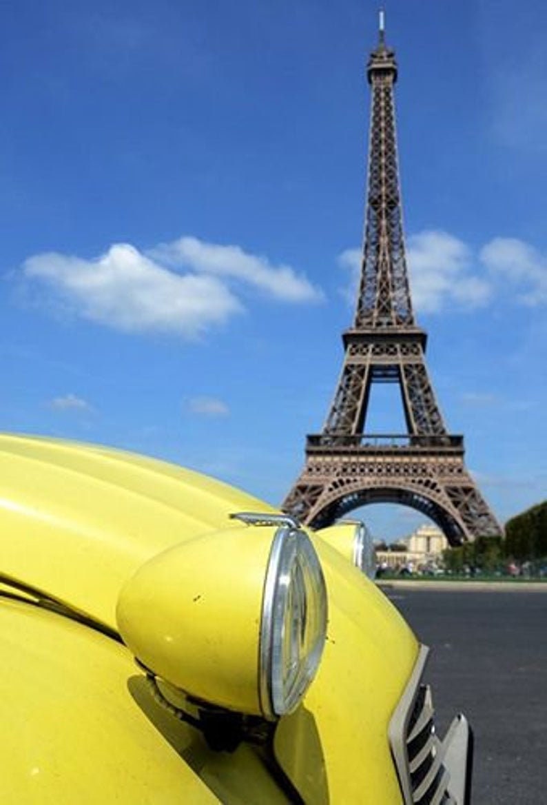 May include: A yellow vintage car with a large headlight is in the foreground, with the Eiffel Tower in the background under a blue sky with clouds. The car's bonnet curves towards the camera.