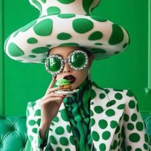May include: A person wearing a white and green polka dot suit with a large green and white polka dot mushroom hat and green rhinestone sunglasses. The person is holding a small green cupcake.