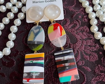 May include: A pair of Marion Godart earrings with a white pearl-like stud, connected to an oval and rectangular pendant. The oval pendants have colorful abstract designs. The rectangular pendants have a layered design with various colors. A pearl necklace is in the background.