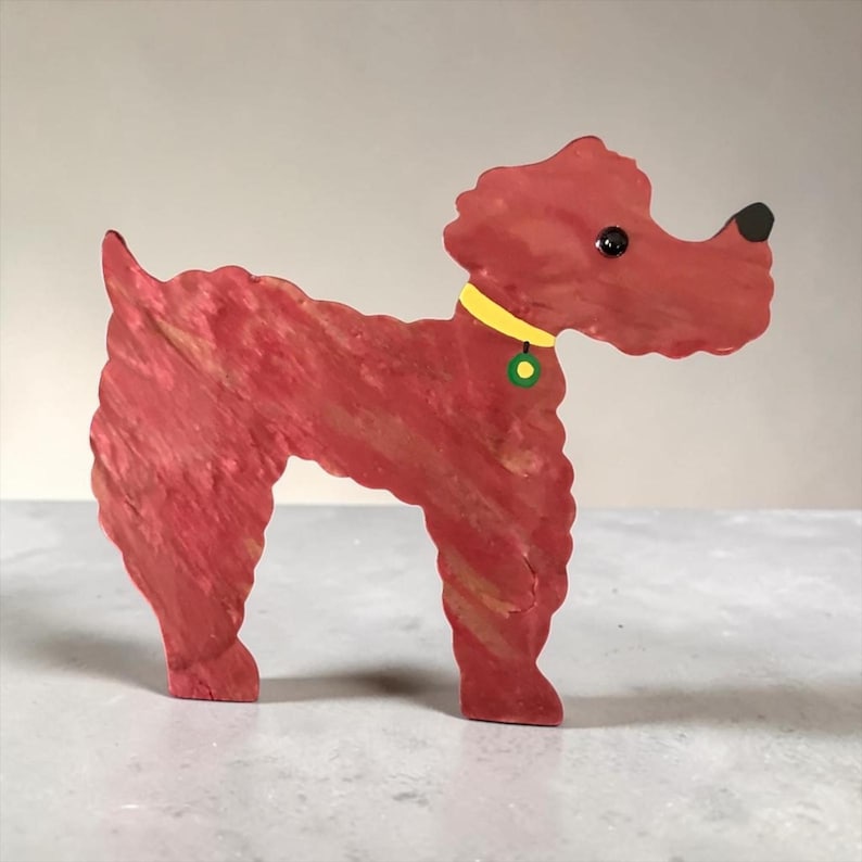 May include: A wooden dog figurine in shades of red with a yellow collar and a green and yellow tag. The dog has a black nose and eye. The figurine is standing on a light gray surface.