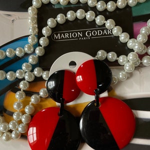 May include: A pair of black and red circular earrings with a contrasting color design. The earrings are on a white card with the text "MARION GODART PARIS". A pearl necklace is in the background.