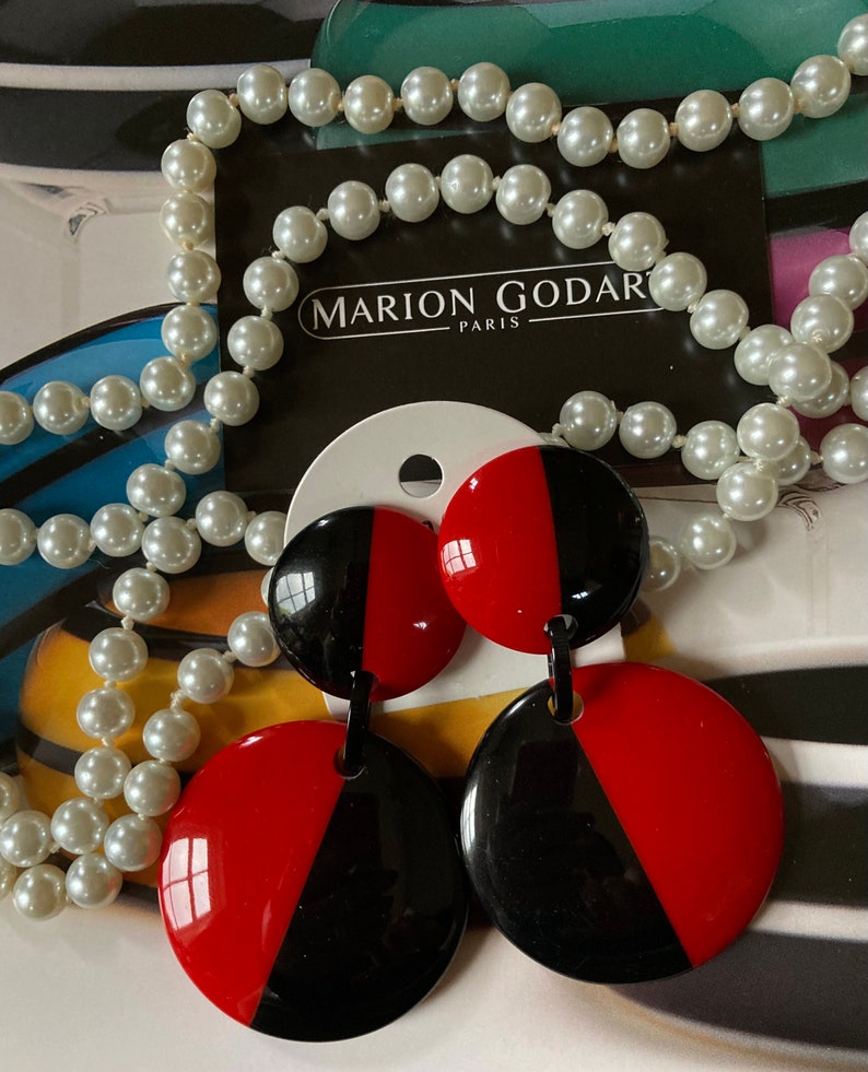 May include: A pair of black and red circular earrings with a contrasting color design. The earrings are on a white card with the text "MARION GODART PARIS". A pearl necklace is in the background.