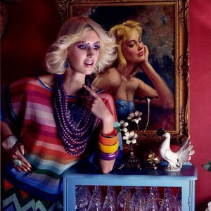 May include: A woman with blonde hair and colorful makeup wears a striped sweater, a purple beaded necklace, and multiple colorful bracelets. She is standing in front of a blue cabinet with glass shelves and a framed painting of a woman with blonde hair.