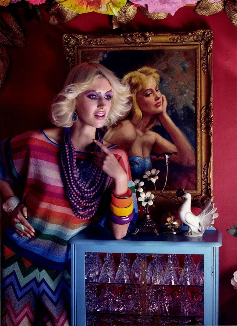 May include: A woman with blonde hair and colorful makeup wears a striped sweater, a purple beaded necklace, and multiple colorful bracelets. She is standing in front of a blue cabinet with glass shelves and a framed painting of a woman with blonde hair.