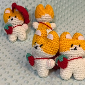 May include: Four crocheted stuffed animals in the shape of foxes. The foxes are white with yellow and red accents. They are holding small red strawberries.