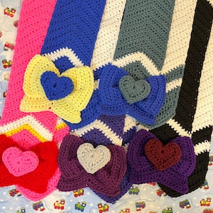 May include: Five crocheted scarves with different colored hearts. The scarves are pink, blue, gray, black, and purple. Each scarf has a heart-shaped design with a different color heart in the center.