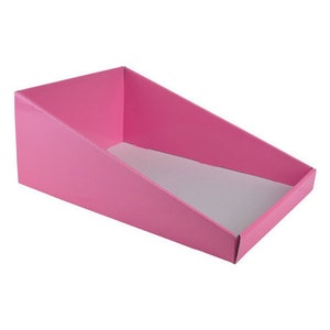 May include: A pink cardboard display stand with a slanted front and a white interior. The stand is designed to hold small items for retail display.