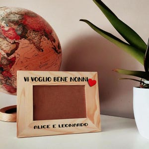 Può includere: Una cornice in legno chiaro con la scritta "VI VOGLIO BENE NONNI" e "ALICE E LEONARDO" in nero. Un piccolo cuore rosso è attaccato alla cornice. Un mappamondo e una pianta in vaso sono sullo sfondo.
