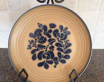 May include: A round ceramic plate with a tan base and a dark blue floral pattern in the center. The plate features concentric ridges along the rim and a dark blue edge. It is displayed on a black metal stand with a decorative scrollwork top.