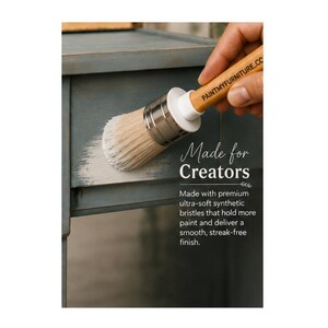 May include: A round paint brush with a light wood handle and white synthetic bristles applying white paint to a blue wooden surface. The handle features the text PAINTMYFURNITURE.COM, and the image includes the text Made for Creators.