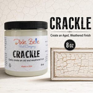 May include: A clear jar of Dixie Belle Paint Company Crackle medium with a black lid and a white label featuring the text CRACKLE, 8 oz, and Made in USA. Next to it is a rectangular frame showing a white surface with a cracked, weathered finish.