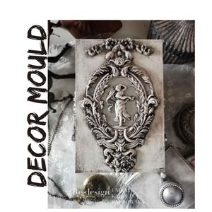 May include: A rectangular, off-white decor mould with a detailed, ornate oval frame. Inside the frame is a relief of a figure with outstretched arms. The frame and figure are in a silver-grey color. The words "DECOR MOULD" are printed vertically in black.