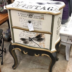 Pode incluir: Uma mesa auxiliar vintage branca com detalhes em preto e dourado. A mesa tem duas gavetas com texto em franc&ecirc;s e um design de lib&eacute;lula. A mesa est&aacute; sobre um piso de cor marrom claro.