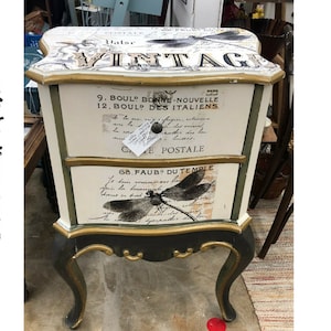 Pode incluir: Uma mesa de cabeceira de estilo vintage com duas gavetas com acabamento pintado em branco e verde. As gavetas s&atilde;o decoradas com papel decoupage com texto franc&ecirc;s e um design de lib&eacute;lula. A mesa de cabeceira tem detalhes dourados e pernas ornamentadas.