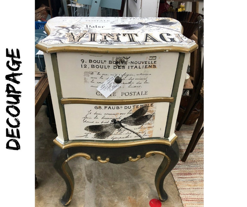 Pode incluir: Uma mesa de cabeceira de estilo vintage com duas gavetas com acabamento pintado em branco e verde. As gavetas s&atilde;o decoradas com papel decoupage com texto franc&ecirc;s e um design de lib&eacute;lula. A mesa de cabeceira tem detalhes dourados e pernas ornamentadas.