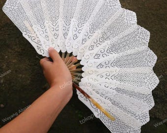 May include: A white paper fan with an elaborate, cut-out design. The fan is held open, revealing its full, semi-circular shape. The intricate patterns are visible across the fan's surface. The handle is made of light-colored wood. The fan is open.