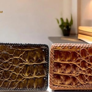 May include: Two leather wallets with a crocodile skin texture. One is dark brown with a black border, and the other is light brown with a braided border. The wallets are rectangular and appear to be new.