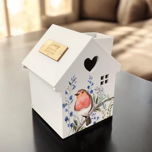 May include: White wooden birdhouse with a robin and blue flowers painted on the side. The roof has a small wooden plaque with the inscription "Nanny, Our Very Own Angel,  September 20, 1937".