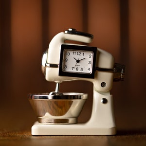May include: White stand mixer clock with a silver bowl and silver clock face. The clock face has black hands and black numbers.