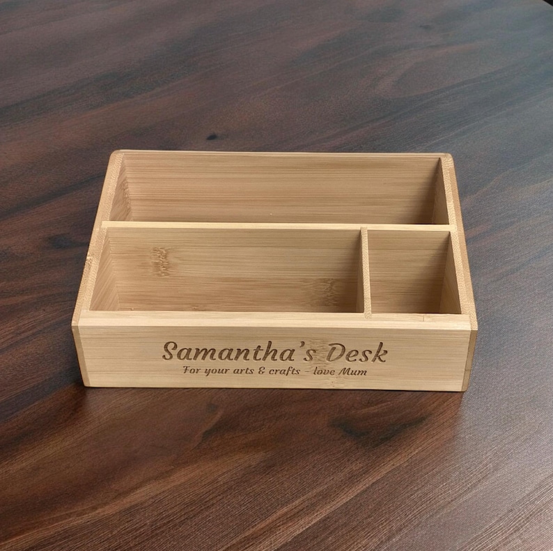 Op de afbeelding: Een lichtgekleurde bamboe bureau-organizer met drie compartimenten. De voorkant van de organizer heeft de tekst "Samantha's Desk" en "For your arts & crafts - love Mum" gegraveerd. De organizer staat op een donkere houten ondergrond.
