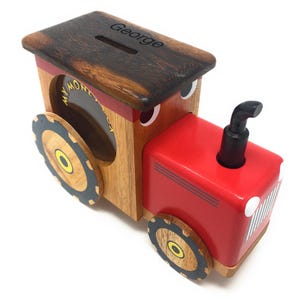 May include: A red wooden toy tractor money box with a slot on the side and a black exhaust pipe. The top of the money box is a wooden lid with the name "George" engraved on it.