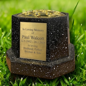 May include: A black and gold memorial vase with a speckled design. The vase features a gold plaque inscribed with "In Loving Memory of Paul Walcott 12.5.1952-28.1.2025 A loving Husband, Father, Brother & Son."