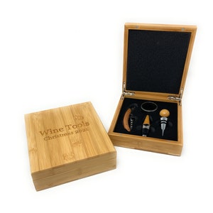 May include: A wooden box with a hinged lid, containing a wine opener, a bottle stopper, and a foil cutter. The box is engraved with the text "Wine Tools Christmas 2021".