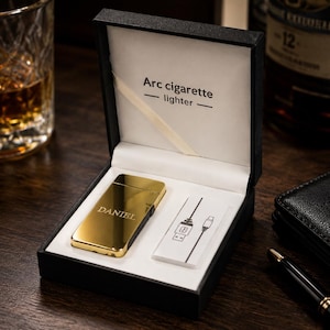 May include: A gold-coloured rectangular arc cigarette lighter with the name DANIEL engraved on the front, presented in a black box with white interior lining. The box lid features the text Arc cigarette lighter and includes a USB charging cable.