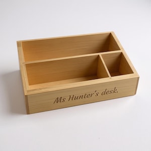 Op de afbeelding: Een lichtbruine houten bureau-organizer met drie compartimenten. De tekst "Ms Hunter's desk." is op de voorkant gegraveerd. De organizer is gemaakt van lichtgekleurd hout.