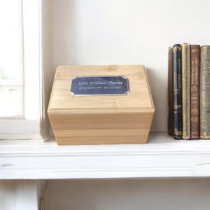 May include: A light brown wooden box with a silver plaque that reads "John William Davies 8th September 1930 - 28th June 2023". The box is sitting on a white shelf with a stack of books behind it.