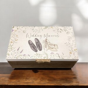 May include: A white wooden box with a floral design and the text "Wedding Memories". The box has a rope handle and features a pair of black shoes and a pair of white shoes.