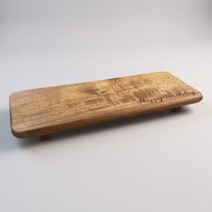 May include: A rectangular wooden serving board with rounded corners and short feet, featuring a natural light brown wood grain pattern. The text "Happy Birthday" is engraved in a dark cursive script on the right side of the board surface.
