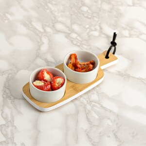 May include: A white ceramic serving tray with two small bowls. The tray is made of bamboo and has a white painted edge. The bowls contain red peppers stuffed with cheese and dried tomato slices.