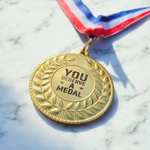 May include: Gold-coloured medal with a laurel wreath design and the text "YOU DESERVE A MEDAL" on the front. The medal is attached to a red, white and blue ribbon.