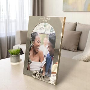 May include: Silver photo frame with an arched opening, featuring a photo of a couple smiling. The frame has an inscription that reads "Jane & Andy - 1.1.2023" and "Amore" with a decorative ring design at the bottom.