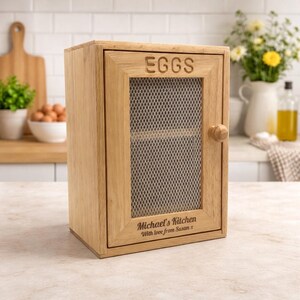 May include: A light brown wooden egg storage cabinet with a hinged door featuring a metal mesh screen. The word EGGS is engraved at the top, and the text Michael's Kitchen With love from Susan x is engraved at the bottom of the door frame.