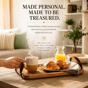 May include: A rectangular acacia wood serving tray with dark brown leather handles. The tray holds a two-toned ceramic mug, a small plate with two croissants, a glass pitcher of orange juice, and a folded linen napkin on a wooden table.