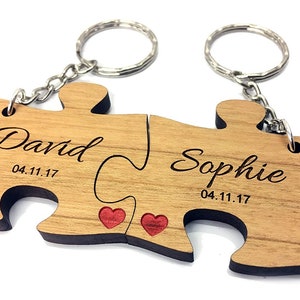 May include: A wooden puzzle keychain with two interlocking pieces, each engraved with a name and date. The names are "David" and "Sophie" with the date "04.11.17" on each piece. The pieces are connected by a silver chain with two key rings.