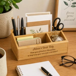 Op de afbeelding: Een bamboe bureau-organizer met meerdere compartimenten, met pennen, notitieblokken en briefpapier. De voorkant van de organizer is gegraveerd met de tekst "Adam's Desk Tidy" en een bericht. Zwarte schaar en plakband zijn ook zichtbaar.