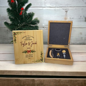 May include: A wooden box with a Christmas-themed design. The box has a hinged lid and contains a bottle opener, a wine stopper, and a foil cutter. The lid has a personalised message that reads "Wishing you a very Merry Christmas Taylor & Jacob 2023 Love always Mum & Dad xxx".