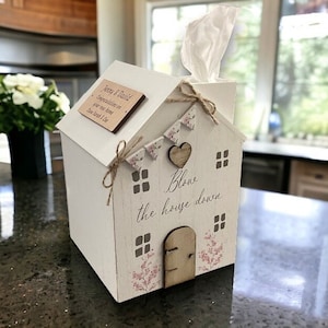 May include: White wooden house-shaped tissue box cover with a pink floral design and the text "Blow the house down". The cover has a small wooden door and a heart-shaped cutout. A personalised wooden plaque with the text "Jenny & David" is attached to the side.