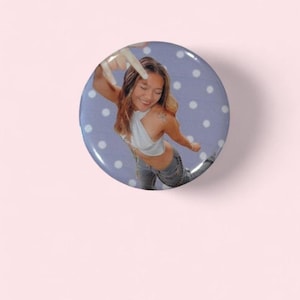 May include: A round button with a light purple background and white polka dots. The button features a person wearing a white top and jeans. The person is making a hand gesture.