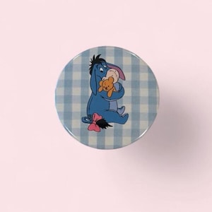 May include: A circular knob featuring a blue and white checkered pattern background. Centered on the knob is a graphic illustration of a blue donkey character holding a small tan bear. The donkey has a pink bow attached to its tail and a pink inner ear.