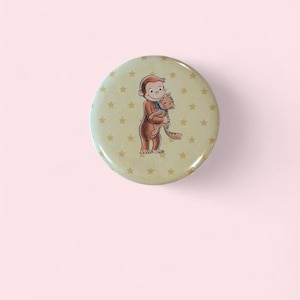 May include: A circular button featuring a light tan background with a repeating pattern of small yellow stars. In the center, a brown monkey character is depicted holding a small orange cat with a blue collar in its arms.