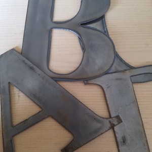 May include: A collection of large, flat, cut-out metal letters in a dark gray, industrial finish. The letters include a prominent capital B, a capital A, and a capital T, all featuring clean, sharp edges and a smooth, matte metallic surface texture.