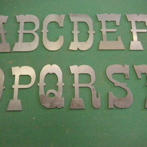 May include: A collection of individual metal letters cut from sheet metal, featuring a distinct western-style serif font. The set includes the uppercase letters A, B, C, D, E, F, G, O, P, Q, R, S, and T, all displaying a raw, unpainted gray finish.