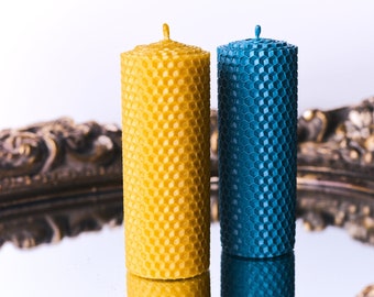 Op de afbeelding: Twee cilindrische kaarsen met een honingraatpatroon. Eén is geel, de andere is blauwgroen. Ze staan op een spiegelend oppervlak, dat hun vormen weerspiegelt. De kaarsen hebben een decoratief, gestructureerd uiterlijk, geschikt voor interieurdecoratie.