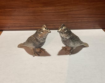 Può includere: Un paio di saliere e pepiere argentate a forma di uccello. Gli uccelli hanno dettagliate trame di piume e sono appollaiati su piccole basi a forma di ramo. Ogni uccello ha una piccola apertura sulla parte superiore.