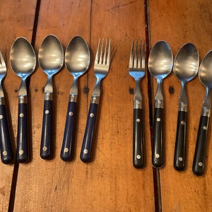 May include: A collection of stainless steel forks and spoons with dark blue and black handles. The silverware is arranged on a wooden surface, showcasing the contrasting colors and textures of the utensils.