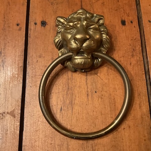 May include: A brass door knocker featuring a sculpted lion head with a detailed mane and facial features. A large, circular brass ring is suspended from the lion's mouth, serving as the striking element for this functional metal hardware piece.